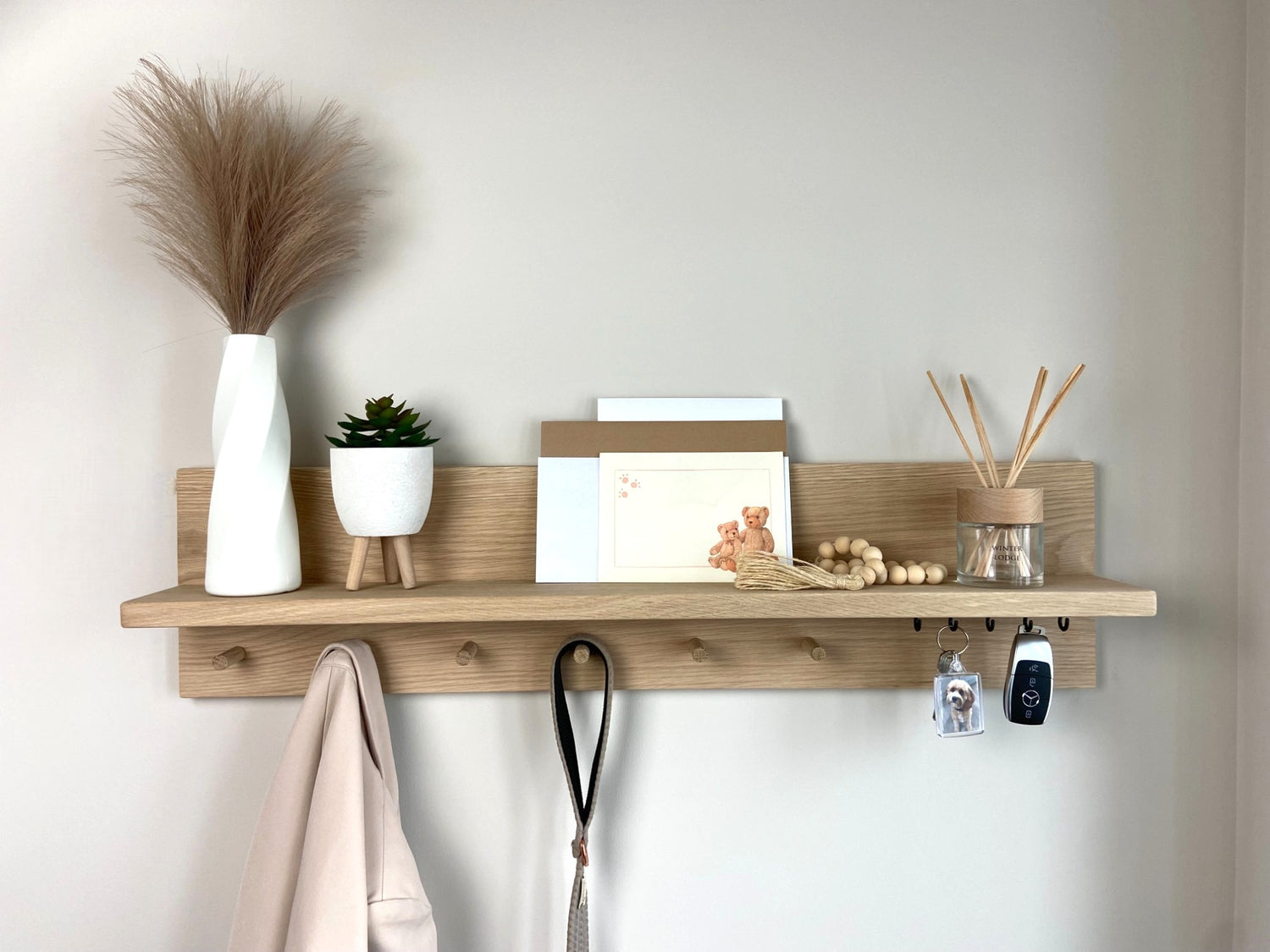 Handcrafted oak organiser shelf with wooden pegs and hooks displaying decorative items, letters, coat and keys. mounted to a wall in an entryway.