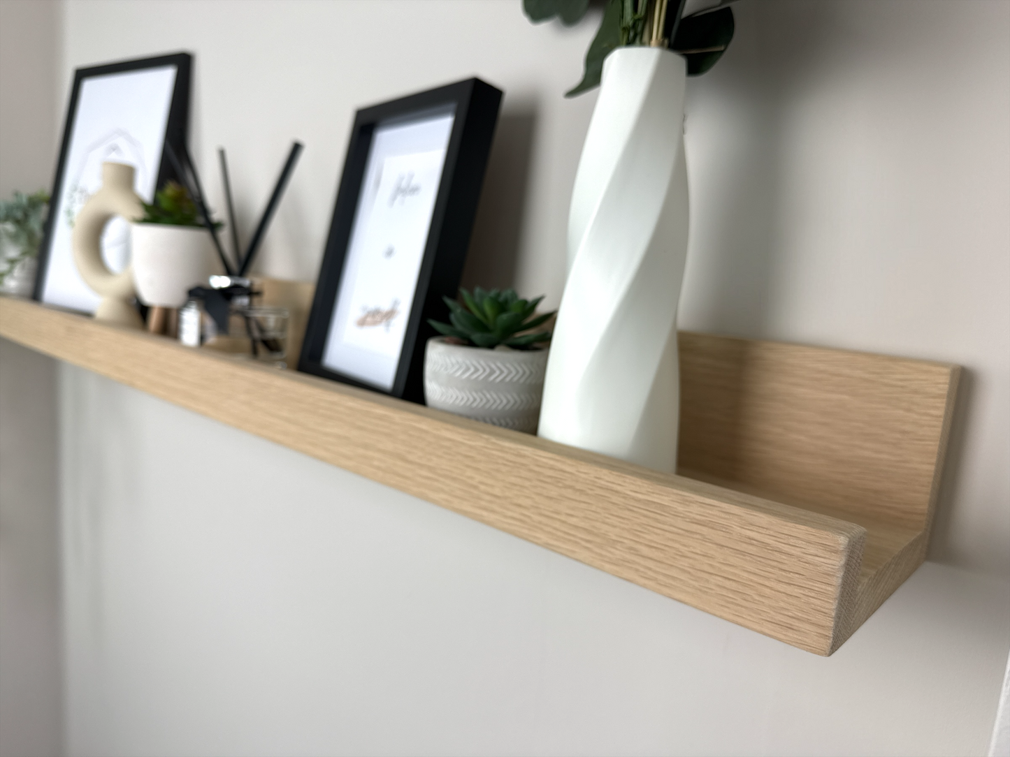 Close-up of the quality craftsmanship of a handmade oak picture shelf, mounted on a plasterboard wall, showcasing a smooth finish and holding photo frames, plants, and small decorative items.